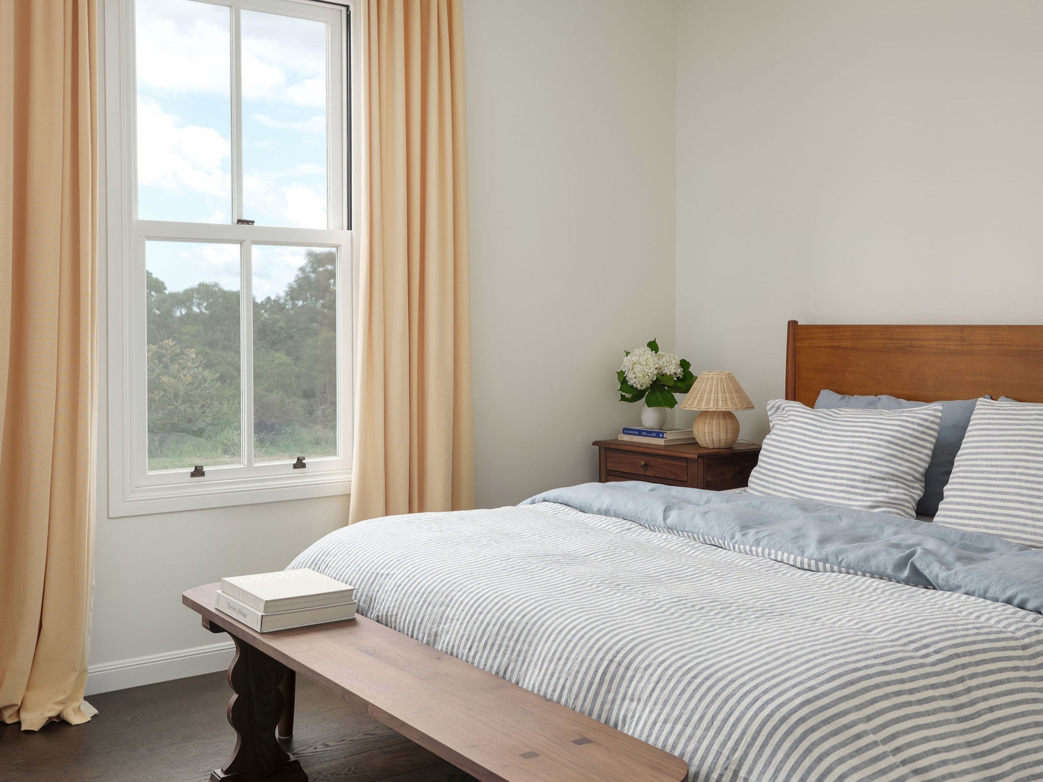 A sunlit guest bedroom with striped bed linen, flowing curtains and walnut finishes. 